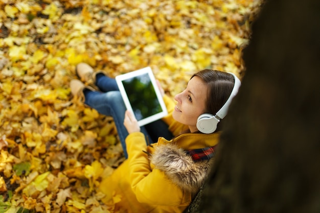 Zdjęcie brązowowłosa kobieta w żółtym płaszczu i dżinsach siedzi i słucha muzyki pod drzewem z tabletem w dłoniach i słuchawkami w jesiennym parku miejskim w ciepły dzień. jesienne złote liście. widok z góry