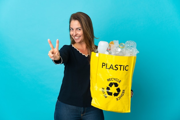 Brazylijka w średnim wieku trzyma torbę pełną plastikowych butelek do recyklingu na niebieskiej ścianie, uśmiechając się i pokazując znak zwycięstwa