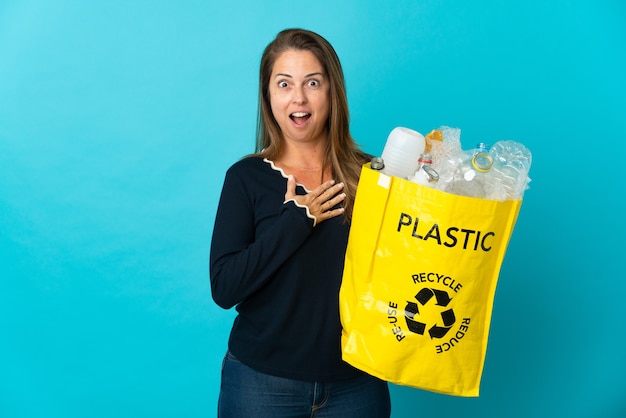 Brazylijka w średnim wieku trzymająca torbę pełną plastikowych butelek do recyklingu na niebieskiej ścianie, zaskoczona i zszokowana, patrząc w prawo