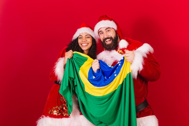 Brazylijska para ubrana na boże narodzenie święty mikołaj mumia mikołaj trzymająca flagę brazylii czarna kobieta i kaukaski mężczyzna