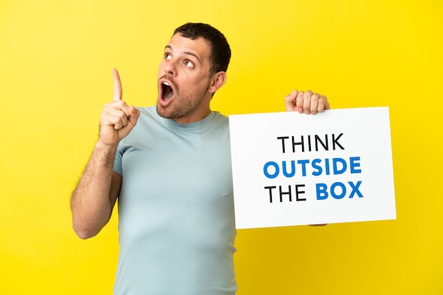 Brazylijski mężczyzna na odosobnionym fioletowym tle, trzymający afisz z tekstem Think Outside The Box and thinking