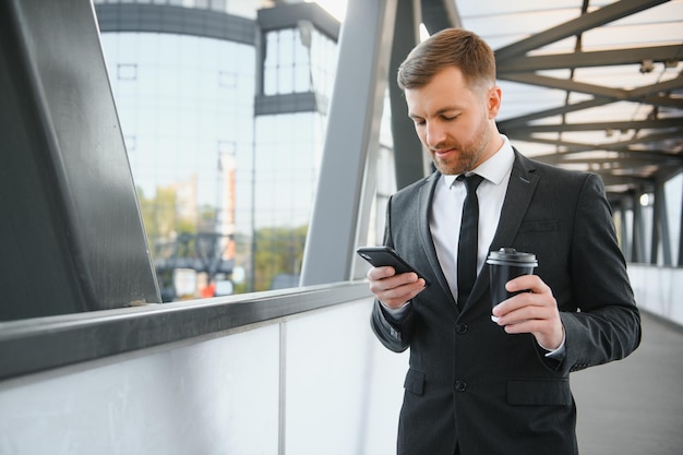 Brodaty biznesmen w formalnym garniturze na przerwie przy użyciu telefonu komórkowego za pomocą smartfona biznes człowiek stojący na zewnątrz na tle ulicy nowoczesnego miasta miejskiego z filiżanką kawy w centrum na zewnątrz miejsca kopiowania