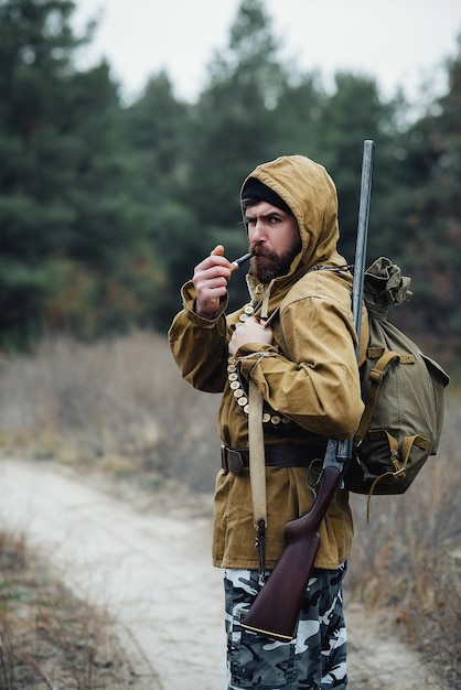 Brodaty Brutalny Myśliwy Mężczyzna W Czarnym Kapeluszu W Kapturze W Kurtce Khaki Z Pistoletem Na Ramieniu I Nabojem Na Piersi Pali Fajkę I Idzie Do Lasu