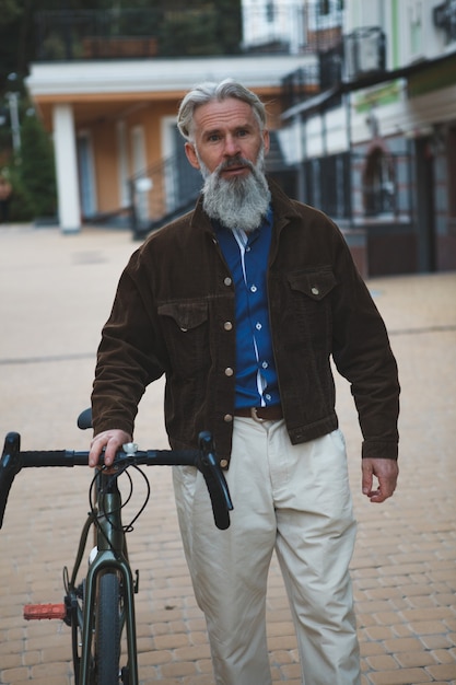 Brodaty dojrzały mężczyzna ubrany w modną kurtkę spacerującą z rowerem