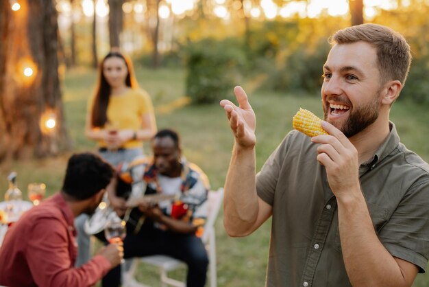 Brodaty hipster jedzący kukurydzę na tle kolacji wielonarodowych przyjaciół