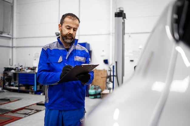 Brodaty kaukaski mechanik samochodowy w średnim wieku robi oględziny pojazdu w warsztacie i trzyma listę kontrolną.