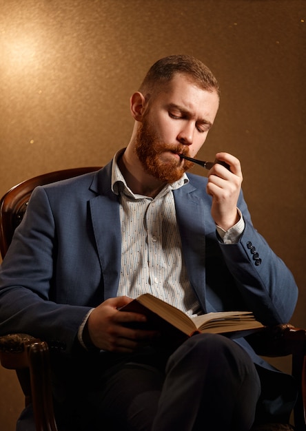 Brodaty Lamber jak męski model w garniturze z wąsami i fajką do brody i czytając książkę, siedząc na krześle.