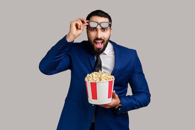 Brodaty mężczyzna jest pod wrażeniem filmu filmowego przytrzymaj popcorn oglądając niesamowity film trzymając otwarte usta podnosząc okulary 3d w oficjalnym stylu garnitur Kryty studio strzał na białym tle na szarym tle