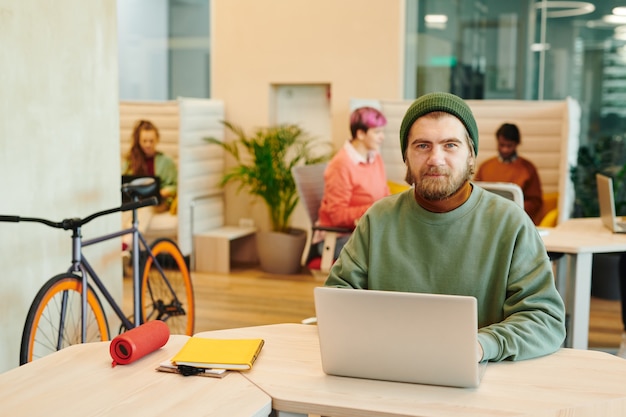 Brodaty mężczyzna kierownik biura w casualwear, patrząc na wyświetlacz laptopa, siedząc w fotelu i przygotowując prezentację przeciwko kolegom