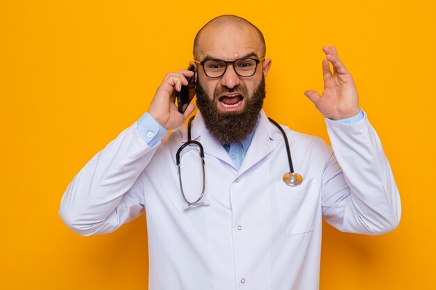 Brodaty mężczyzna lekarz w białym fartuchu ze stetoskopem na szyi w okularach krzyczących z agresywną miną podczas rozmowy przez telefon komórkowy mobile
