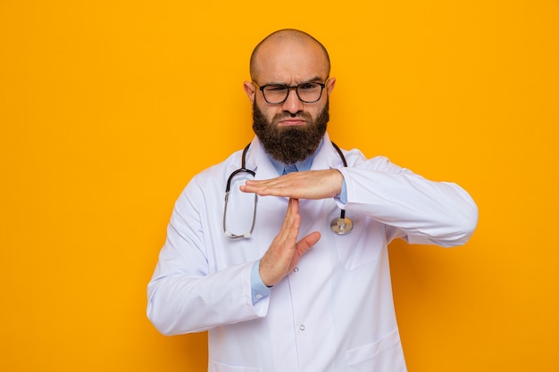 Zdjęcie brodaty mężczyzna lekarz w białym fartuchu ze stetoskopem wokół szyi w okularach, patrząc na kamerę, co limit czasu gest z rękami stojącymi na pomarańczowym tle