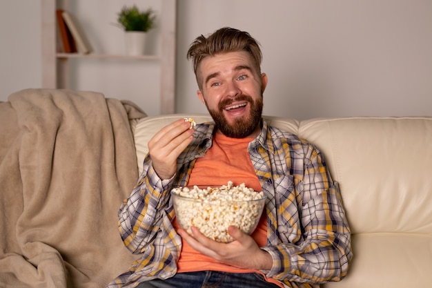 Brodaty mężczyzna ogląda telewizję jedzenie popcornu w domu w nocy.