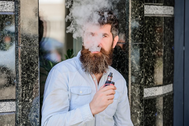 Brodaty mężczyzna palący vape Mężczyzna z brodą wydychający dym Chmury aromatyzowanego dymu Palenie elektronicznego papierosa Koncepcja łagodzenia stresu Urządzenie do palenia Mężczyzna długa broda zrelaksowany z nałogiem palenia