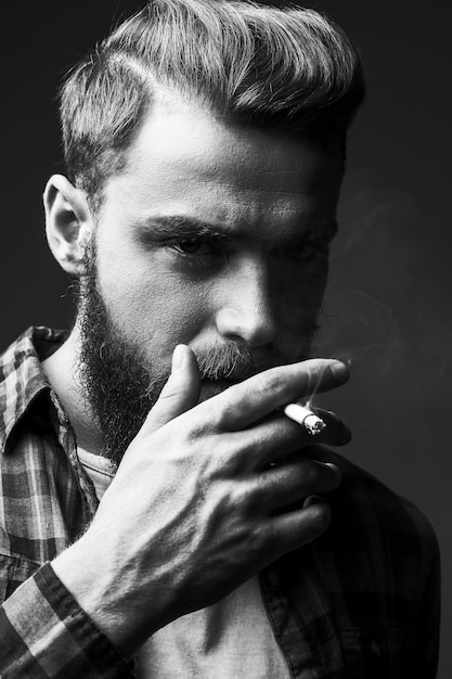 Brodaty mężczyzna palenia. Czarno-biały portret przystojny młody brodaty mężczyzna palący papierosa i patrzący na kamerę stojąc na szarym tle