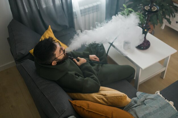 Brodaty mężczyzna pali fajkę wodną w domu i wydmuchuje chmurę dymu w czasie chłodu i koncepcji odpoczynku