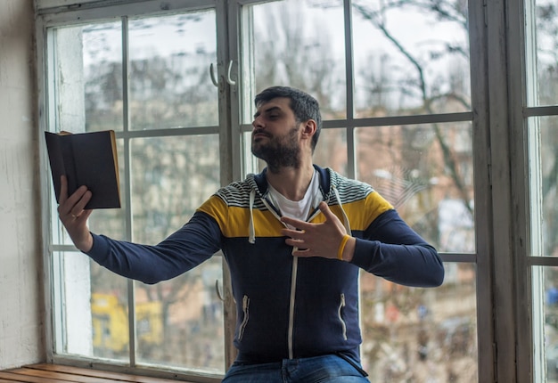 Zdjęcie brodaty mężczyzna poeta czyta książkę z gestem ekspresji. duże okno na tle.
