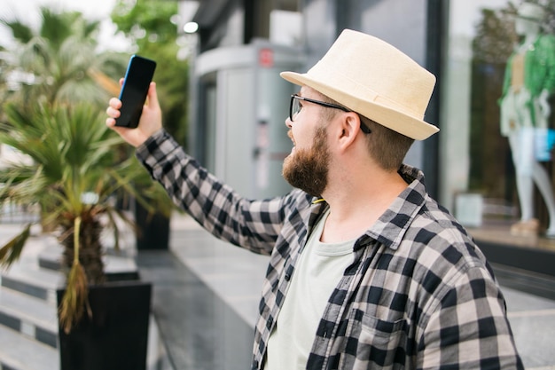 Brodaty mężczyzna robi selfie za pomocą smartfona przechadza się po śródmieściu pozuje na tle nowoczesnego budynku miejskiego cieszy się wolnym czasem Ludzie miejski styl życia i koncepcja technologii