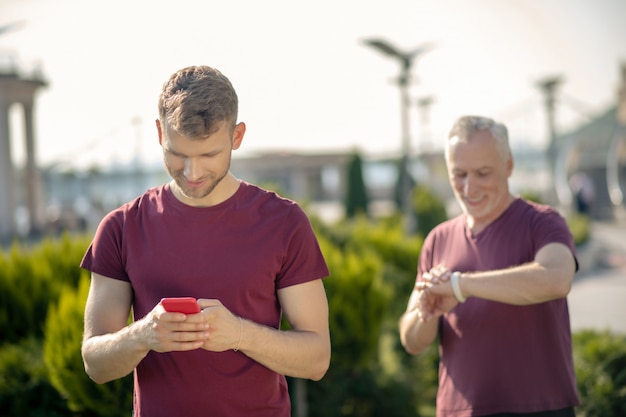 Brodaty Mężczyzna Sprawdzający Swój Smartfon, Siwy Mężczyzna Sprawdzający Smartwatch Za Nim