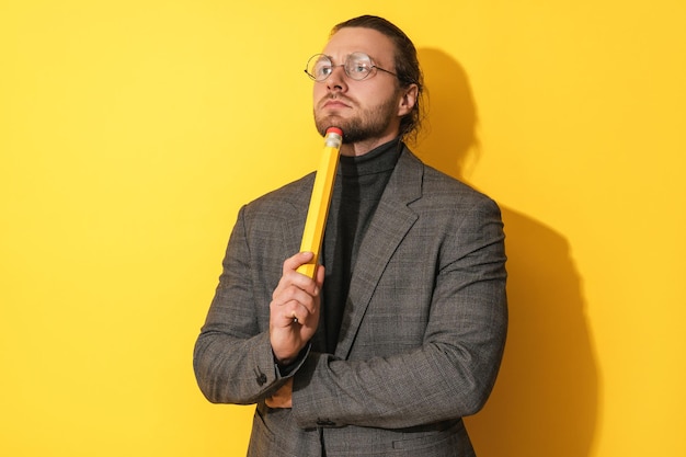 Brodaty mężczyzna w okularach trzymający duży ołówek i notatnik zaskoczony na żółtym tle