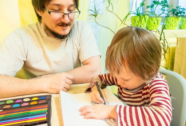 Brodaty mężczyzna w szarej koszulce i słodkim chłopcu rysującym obrazki kolorowymi kredkami woskowymi podczas wspólnego spędzania czasu w domu