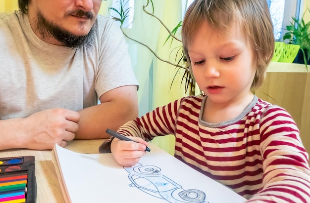 Brodaty mężczyzna w szarej koszulce i słodkim chłopcu rysującym obrazki kolorowymi kredkami woskowymi podczas wspólnego spędzania czasu w domu