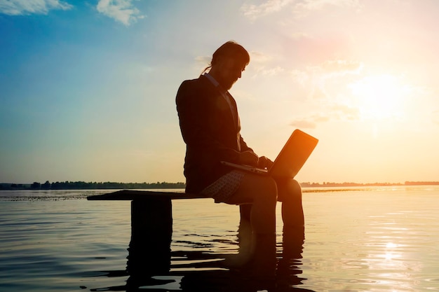 Brodaty mężczyzna w szarej kurtce i bieliźnie siedzi na drewnianej ławce w wodzie z laptopem o zachodzie lub wschodzie słońca