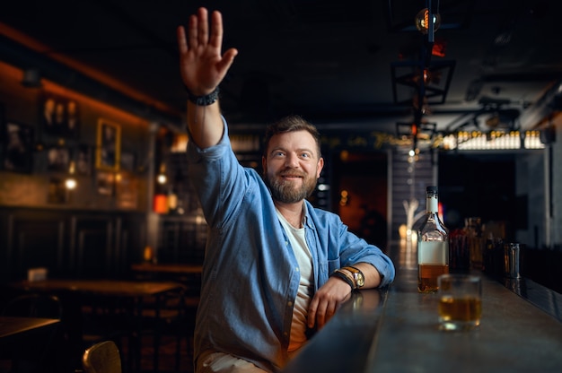 Brodaty mężczyzna z podniesioną ręką siedzi przy kasie w barze. Jeden mężczyzna odpoczywający w pubie, ludzkie emocje, zajęcia rekreacyjne, życie nocne