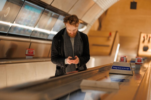 Brodaty mężczyzna za pomocą smartfona stojąc na schodach ruchomych w metrze
