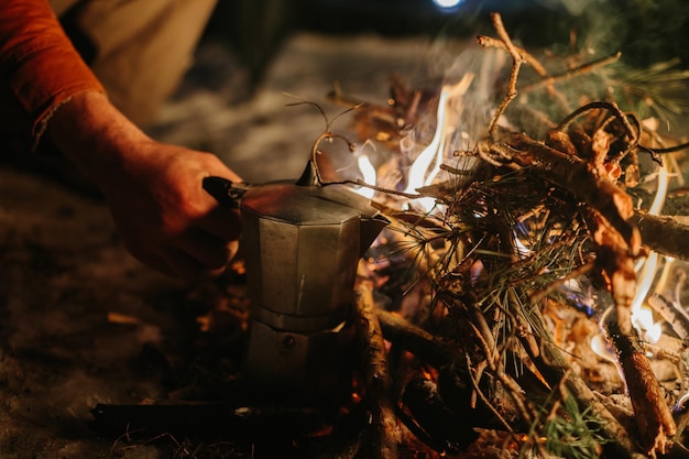Brodaty młody człowiek robi pyszną kawę na ognisku, podziwiając zachód słońca w zimowym lesie Koncepcja przetrwania i wędrówki w dziczy