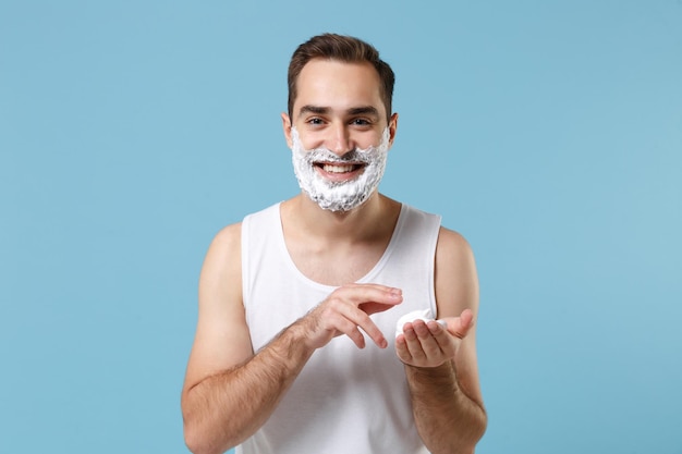 Brodaty młody mężczyzna w wieku 20 lat w białej koszuli twarz pokryta pianką do golenia odizolowany na niebieskim pastelowym tle portretu studyjnego. Koncepcja zabiegów kosmetycznych do pielęgnacji skóry. Makieta miejsca na kopię.
