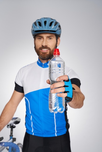 Zdjęcie brodaty rowerzysta w kasku pokazujący butelkę wody do aparatu w pomieszczeniu, portret cyklu