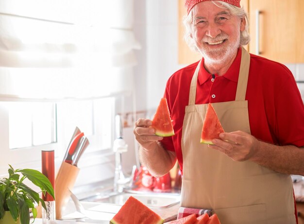 Brodaty starszy mężczyzna z białymi włosami i czerwoną chustką trzymający dwa plasterki deski do krojenia arbuza i przybory kuchenne na stole aktywni starsi ludzie bawią się w kuchni