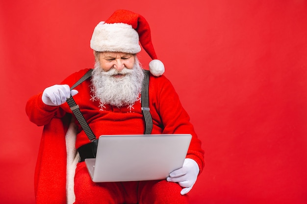 brodaty starzec w santa costime trzymając laptopa na białym tle na czerwonym tle