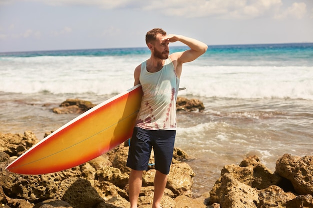 Brodaty Surfer Z Deską Surfingową Spaceruje Po Plaży I Na Miejscu Wypatruje Fal