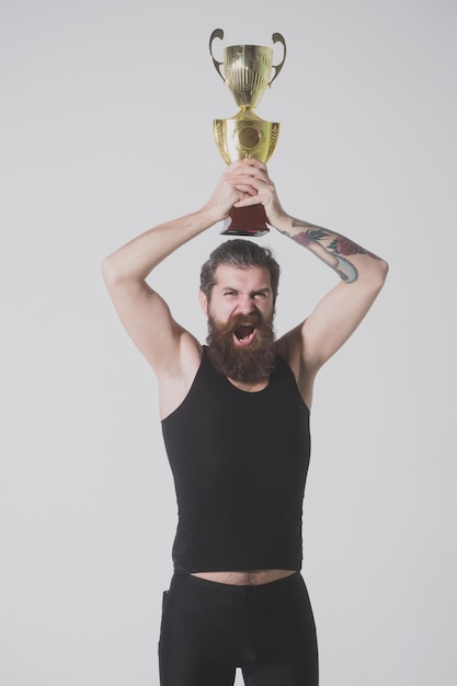 Zdjęcie brodaty szczęśliwy mężczyzna trzyma złoty puchar mistrza na szarym tle