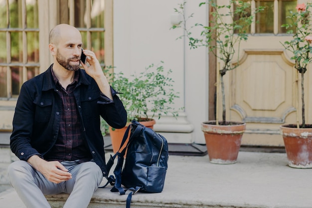 Brodaty turysta z gęstą brodą rozmawia przez telefon odpoczywa po wycieczce używa nowoczesnego telefonu komórkowego do komunikacji zamyślony patrzy na bok podziwia cudowną scenerię Koncepcja technologiczna