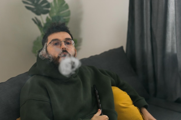 Brodaty tysiącletni mężczyzna lub gen z palący fajkę wodną podczas relaksu na kanapie w domu i odpoczynku