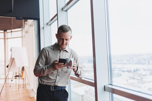 Brokerzy płci męskiej z telefonem patrzącym w stronę okna z kawą w ręku w nowoczesnym miejscu pracy
