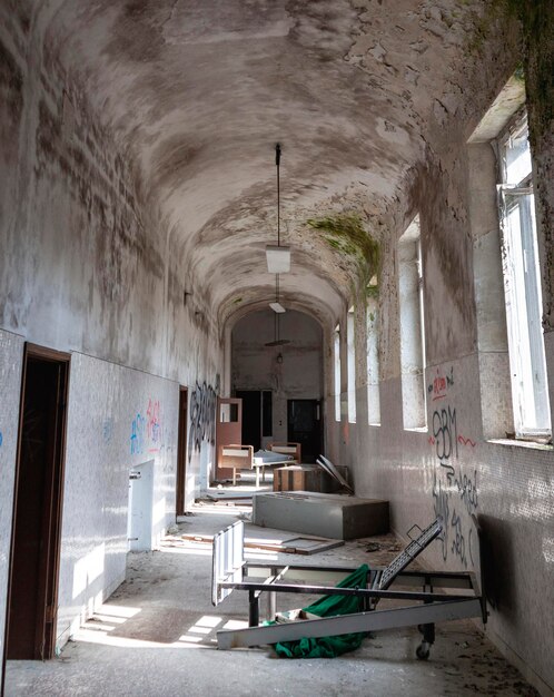 Brudny, zniszczony betonowy korytarz w starym opuszczonym budynku. Na korytarzu porozrzucane są sprzęty, meble i łóżka.