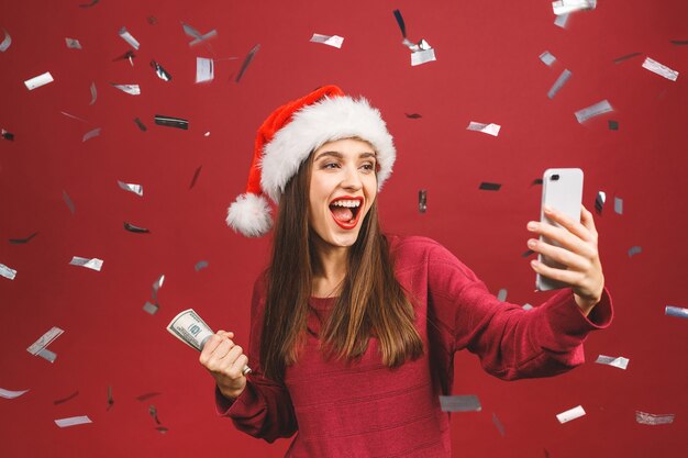 Brunetka dama w kapeluszu Boże Narodzenie, trzymając pieniądze i biorąc koncepcja selfie