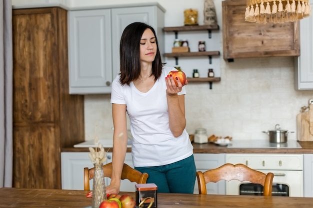 Brunetka dziewczyna w białej koszulce w kuchni je jabłko. Wysokiej jakości zdjęcie