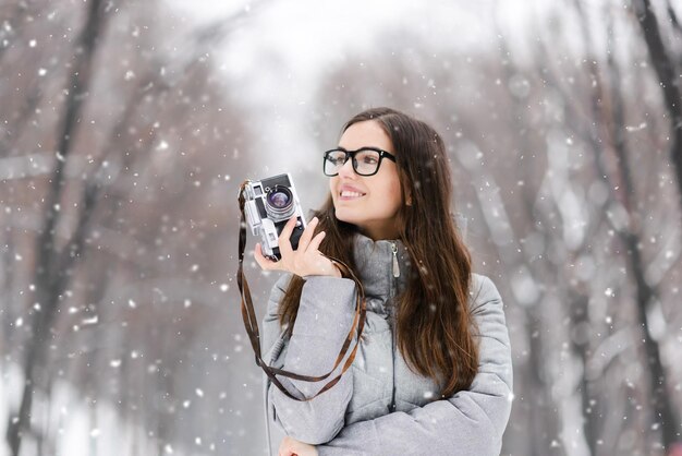 Brunetka fotograficzna dziewczyna w okularach ubrana w szarą kurtkę, patrząca w górę i trzymająca retro aparat w zimowym lesie