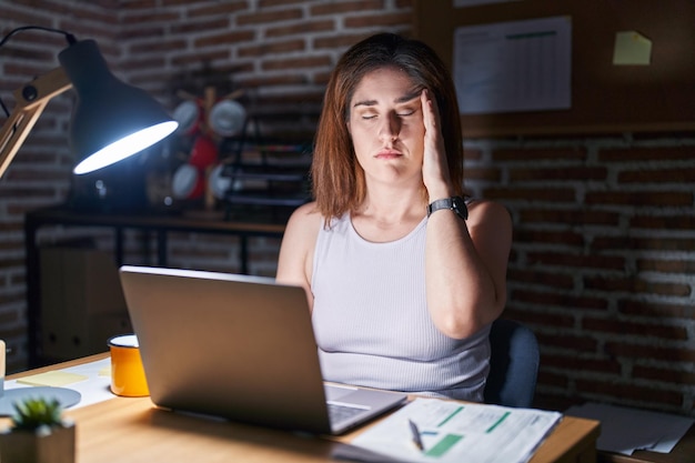 Brunetka kobieta pracująca w biurze w nocy z ręką na głowie z powodu bólu głowy z powodu stresu cierpiącego na migrenę