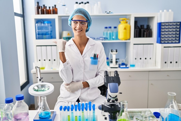 Brunetka kobieta pracująca w laboratorium naukowca uśmiecha się z radosną twarzą, patrząc i wskazując na bok kciukiem do góry