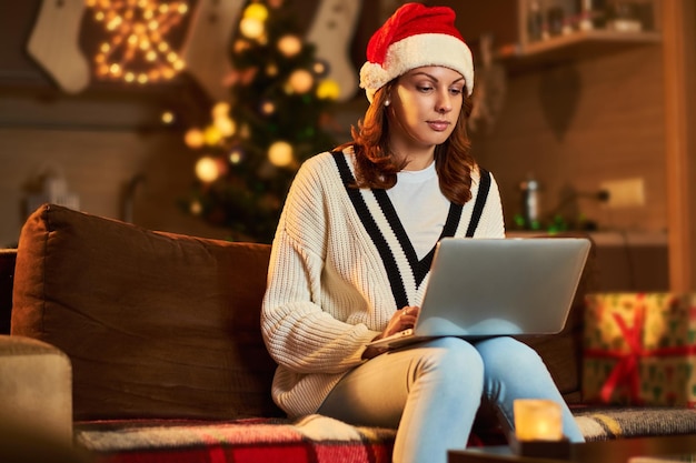 Brunetka Kobieta W Kapeluszu świętego Mikołaja Za Pomocą Laptopa W świątecznej Dekoracji Salonu.