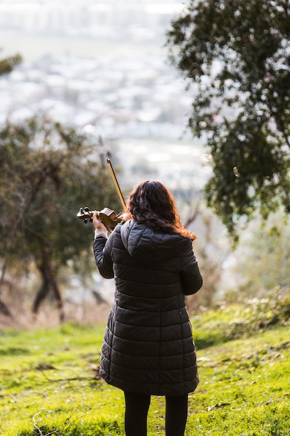 Brunetka kobieta widziana z tyłu gry na skrzypcach na zewnątrz w górach. Pionowy