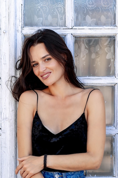 Brunetka ładny model pozuje na tle okna. Nagie ramiona. Czarny top. Ładny uśmiech. Portret do połowy długości.