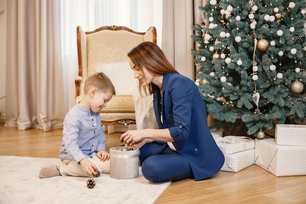 Brunetka Matka I Synek Siedzi Na Podłodze W Pobliżu Choinki W Domu. Rodzina Udekoruje Choinkę Razem Z Bombkami. Kobieta Ubrana W Niebieski Kostium I Niebieską Koszulę Chłopca.