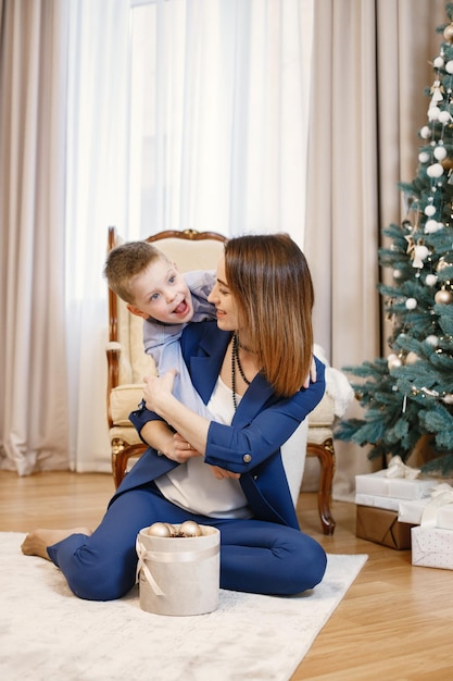 Brunetka matka i synek ze śmiesznymi twarzami siedzą na podłodze w pobliżu choinki w domu. Rodzina udekoruje choinkę razem z bombkami. Kobieta ubrana w niebieski kostium i chłopiec blu