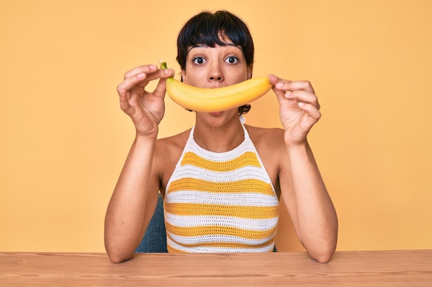 Brunetka nastolatka trzymająca banana jak śmieszny uśmiech przestraszona i zszokowana zaskoczeniem i zdumionym wyrazem strachu i podekscytowaną twarzą